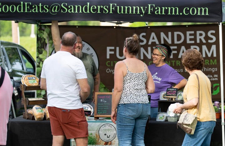 A pop-up canopy tent at an outdoor farmers market or fair, with several people standing around a table. The black canopy has green text reading 'GoodEats@SandersFunnyFarm.